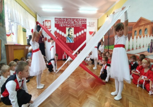 Dziewczynki w białych sukienkach z czerwonymi paskami oraz chłopcy w białych koszulach, czerwono-czarnych kamizelkach oraz czarnych spodniach tańczą z biało-czerwonymi szarfami.